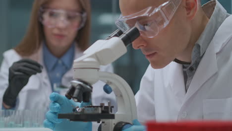 Dos-Técnicos-Están-Trabajando-En-Un-Laboratorio-Científico,-Un-Hombre-Está-Investigando-Muestras-Con-Un-Microscopio-Y-Una-Mujer-Trabajando-Con-Productos-Químicos-En-El-Departamento-De-Exploración-Microbiológica