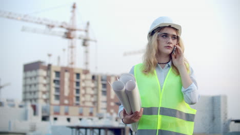 Woman-Builder-talking-on-the-phone-with-the-contractor-with-drawings-in-his-hands-on-the-background-of-buildings-under-construction-and-cranes
