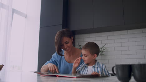 Young-beautiful-mother-and-son-sitting-at-a-table-in-a-bright-kitchen-reading-a-book-and-looking-at-pictures-poking-a-finger-into-a-book-and-leafing-through-the-pages