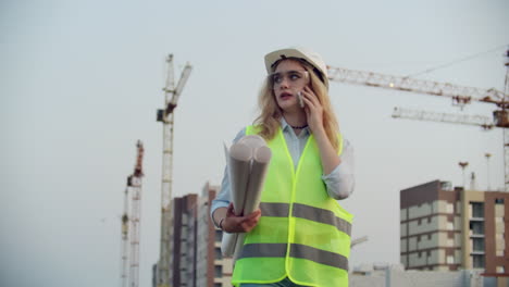 Woman-Builder-talking-on-the-phone-with-the-contractor-with-drawings-in-his-hands-on-the-background-of-buildings-under-construction-and-cranes