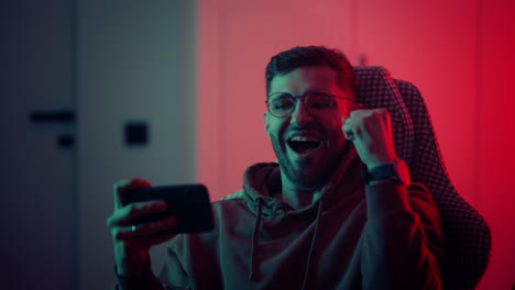 A-young-man-with-glasses-a-professional-gamer-on-a-mobile-phone-plays-games-and-emotionally-rejoices-in-the-victory.-Emotional-young-man-playing-mobile-games-in-a-neon-light.-High-quality-4k-footage