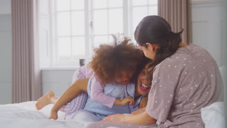 Family-With-Two-Mums-Wearing-Pyjamas-Playing-On-Bed-At-Home-With-Daughter