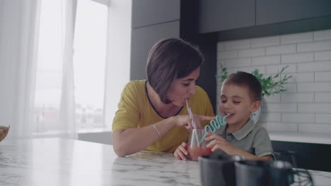 Loving-beautiful-mother-and-son-together-smiling-drinking-orange-juice-from-a-glass-glass-through-a-tube