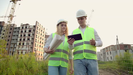 Two-industrial-engineer-wear-safety-helmet-and-holding-tablet-engineering-working-and-talking-with-drawings-inspection.-On-building-outside.-Engineering-tools.