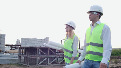 Man-and-woman-supervisor-and-assistant-with-drawings-in-white-helmets-go-and-talk-on-the-construction-site-showing-the-location-of-objects.