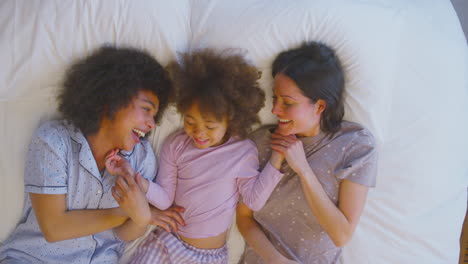 Overhead-Portrait-Of-Family-With-Two-Mums-Wearing-Pyjamas-Playing-On-Bed-At-Home-With-Daughter