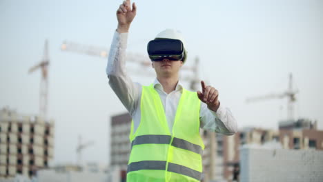 Portrait-of-a-male-inspector-analyzing-the-work-of-builders-using-virtual-reality-glasses.-A-man-in-a-helmet-and-a-protective-vest-stands-in-VR-glasses-and-moves-his-hands.