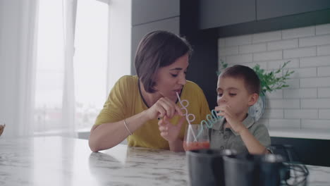 Young-mom-and-son-drinking-juice-with-candy-at-kitchen.-Happy-mother-spending-time-with-son-sitting-on-table-in-slow-motion
