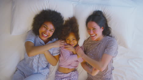 Overhead-Portrait-Of-Family-With-Two-Mums-Wearing-Pyjamas-Playing-On-Bed-At-Home-With-Daughter