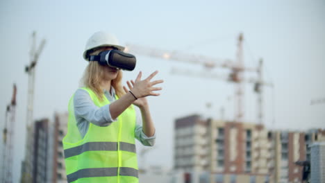 Woman-designer-on-a-building-site-in-hard-hat-and-vest-in-the-glasses-of-virtual-reality-to-move-your-hands-mimicking-the-interface-on-the-background-of-the-cranes-at-sunset.