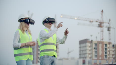 Two-people-in-virtual-reality-glasses-on-the-background-of-buildings-under-construction-with-cranes-imitate-the-work-of-the-interface-for-the-control-and-management-of-construction.