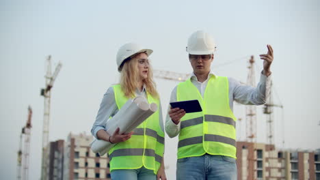 Business-building-industry-technology-and-people-concept---smiling-builder-in-hardhat-with-tablet-pc-computer-along-with-woman-with-drawings-of-builders-at-construction-site