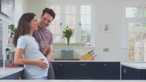 Pregnant-Transgender-Couple-At-Home-In-Kitchen-Relaxing-With-Coffee-As-Man-Touches-Woman's-Stomach