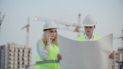 Woman-talking-on-the-phone-and-asks-the-Builder-what-is-on-the-drawings-standing-on-the-background-of-buildings-under-construction.