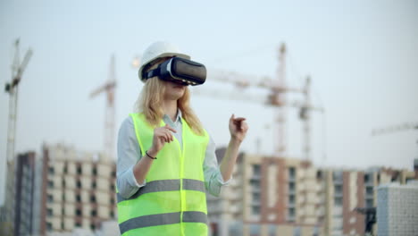 Portrait-of-a-female-inspector-analyzing-the-work-of-builders-using-virtual-reality-glasses.-A-woman-in-a-helmet-and-a-protective-vest-stands-in-VR-glasses-and-moves-his-hands