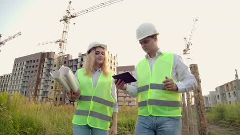 A-man-and-a-woman-in-protective-clothing-and-helmets-go-to-the-construction-site-to-discuss-and-look-around-click-on-the-tablet-screen-on-the-background-of-working-cranes.