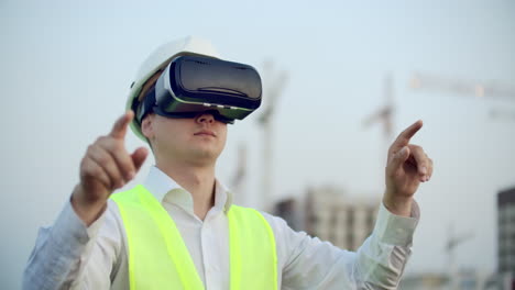 A-man-in-virtual-reality-glasses-helmet-and-vest-on-the-background-of-construction-controls-the-hands-of-the-interface-and-checks-the-quality-of-construction-and-development-of-the-project-and-the-development-plan-and-landscape.-Landscape-designer-uses-virtual-reality.