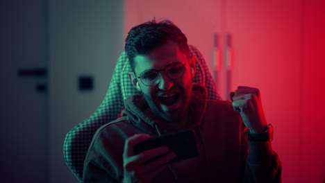 A-young-man-with-glasses-a-professional-gamer-on-a-mobile-phone-plays-games-and-emotionally-rejoices-in-the-victory.-Emotional-young-man-playing-mobile-games-in-a-neon-light.-High-quality-4k-footage