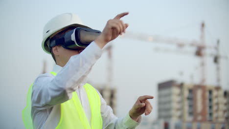 Portrait-of-a-male-inspector-analyzing-the-work-of-builders-using-virtual-reality-glasses.-A-man-in-a-helmet-and-a-protective-vest-stands-in-VR-glasses-and-moves-his-hands.