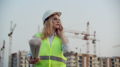 Portrait-of-a-woman-engineer-in-a-helmet-talking-on-the-phone-on-the-background-of-construction-with-cranes-holding-drawings-in-his-hands.-Female-engineer-on-construction-site.