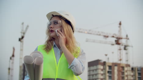 Woman-engineer-designer-talking-on-the-phone-with-the-contractor-with-drawings-in-hand-on-the-background-of-buildings-under-construction-and-cranes