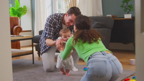 Transgender-Family-Encouraging-Baby-To-Stand-And-Take-First-Steps-In-Lounge-At-Home