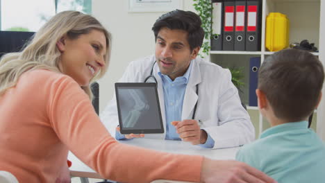 Doctor-Or-GP-In-White-Coat-Meeting-Mother-And-Son-For-Appointment-Looking-At-Scan-On-Digital-Tablet