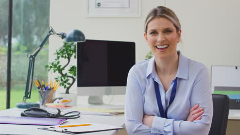 Portrait-Of-Smiling-Female-Doctor-Or-GP-In-Office-Sitting-At-Desk-With-Folded-Arms