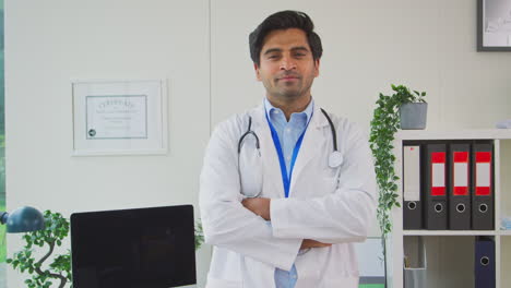 Portrait-Of-Smiling-Male-Doctor-Or-GP-With-Stethoscope-Wearing-White-Coat-Standing-In-Office