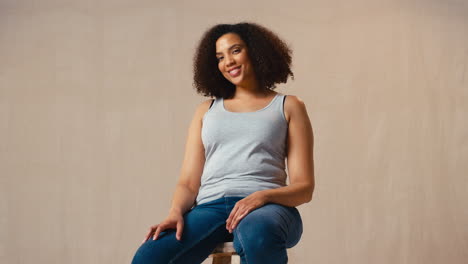 Studio-Portrait-Shot-Of-Casually-Dressed-Body-Positive-Woman-Sitting-On-Stool