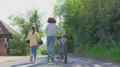 Vista-Trasera-De-Un-Niño-Con-Bicicleta-Y-Una-Niña-Con-Patineta-Caminando-Juntos-Por-Un-Camino-Rural