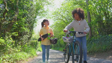 Boy-With-Bike-And-Girl-With-Skateboard-Walking-Along-Country-Road-Together