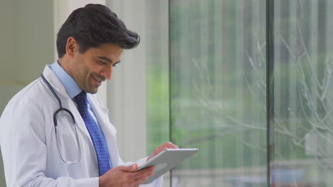 Male-Doctor-Wearing-White-Coat-Standing-In-Hospital-Corridor-Looking-At-Digital-Tablet