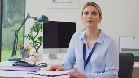 Portrait-Of-Woman-Doctor-Or-GP-In-Office-Sitting-At-Desk-Writing-Notes-On-Clipboard