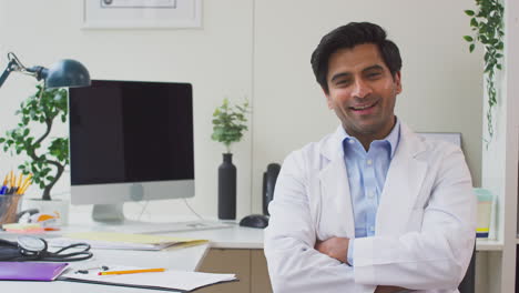 Portrait-Of-Smiling-Male-Doctor-Wearing-White-Coat-Sitting-At-Desk-In-Office