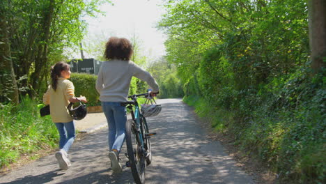 Rückansicht-Eines-Jungen-Mit-Fahrrad-Und-Eines-Mädchens-Mit-Skateboard,-Die-Gemeinsam-Die-Landstraße-Entlang-Gehen