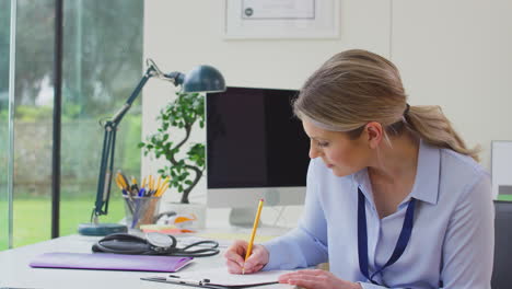 Woman-Doctor-Or-GP-In-Office-Sitting-At-Desk-Writing-Notes-On-Clipboard