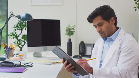 Male-Doctor-Or-GP-Wearing-White-Coat-Sitting-At-Desk-In-Office-Writing-Notes-On-Clipboard