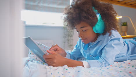 Junge-Im-Schlafzimmer-Liegt-Auf-Dem-Bett-Und-Trägt-Kopfhörer-Und-Nutzt-Ein-Digitales-Tablet