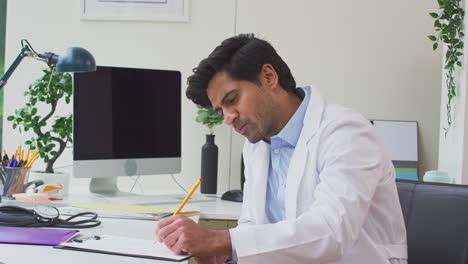 Portrait-Of-Male-Doctor-Wearing-White-Coat-Sitting-At-Desk-In-Office-Writing-Notes-On-Clipboard