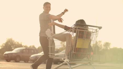 Side-view-of-a-young-man-and-woman-having-fun-outdoors-on-shopping-trolleys.-Multiethnic-young-people-racing-on-shopping-carts.-On-the-parking-zone-with-their