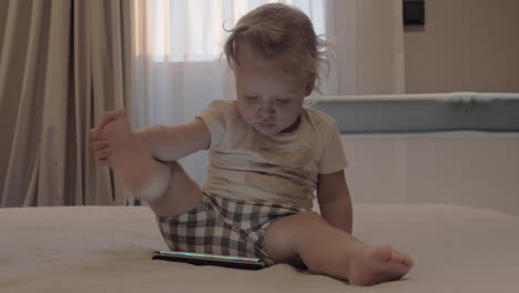 A-baby-girl-in-checkered-shorts-sitting-on-a-bed-with-a-tablet-in-front-of-her