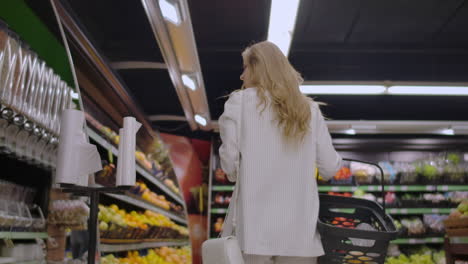 View-back-girl-walking-through-the-supermarket-with-a-basket-in-his-hands-considering-the-shelves-of-fruit