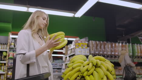 Junges,-Schönes,-Brünettes-Mädchen-In-Ihren-Zwanzigern,-Das-Bananen-Auswählt-Und-Sie-In-Den-Einkaufswagen-In-Der-Obst--Und-Gemüseabteilung-Eines-Lebensmittelladens-Legt