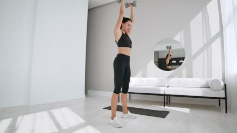 A-young-brunette-girl-in-black-sportswear-performs-squats-with-dumbbells-in-a-white-apartment-on-a-Sunny-day.-Slow-motion