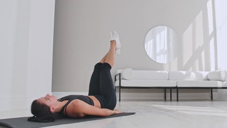 Sportswoman-training-at-home.-Fit-female-athlete-doing-toe-touch-single-arm-exercise-lying-on-floor-in-white-apartment.