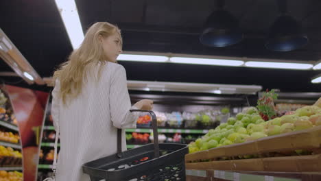 View-back-girl-walking-through-the-supermarket-with-a-basket-in-his-hands-considering-the-shelves-of-fruit