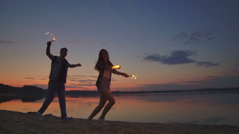 Young-lovers-man-and-woman-cheerfully-run-along-the-beach-with-hot-sparkling-rot-in-their-hands-in-slow-motion.-Romantic-relationship.-Sparklers-burn-in-the-hands-of-lovers