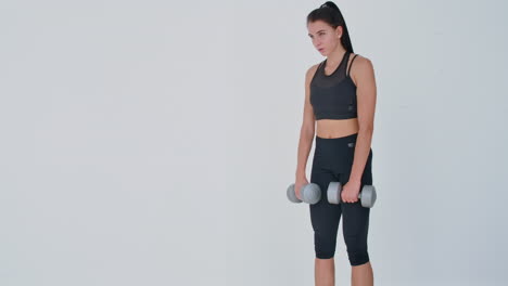 The-beautiful-female-performs-leans-forward-with-dumbbells-in-her-hands-in-the-white-interior-of-the-living-room.-In-slow-motion-to-do-exercises-on-the-muscles-of-the-thighs