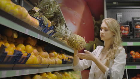 Frau-Wählt-Eine-Ananas-Aus-Und-Legt-Sie-In-Einen-Einkaufskorb,-Nimmt-Eine-Kleine-Ananas,-Die-Daneben-Liegt.-Kundin-In-Der-Obst--Und-Gemüseabteilung-Eines-Großen-Modernen-Supermarkts